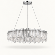 A modern circular chandelier with a polished silver-tone metal frame and numerous clear crystal prisms and hanging drops, suspended by thin cables from a round ceiling canopy against a light off-white background.