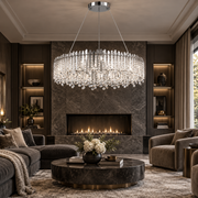 A large circular crystal chandelier with a silver-toned frame hangs over a modern fireplace in a luxurious living room with dark wood paneling, plush seating, and a round marble coffee table.